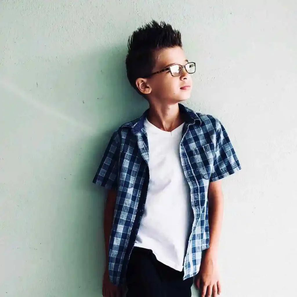 Boy in a white t-shirt and plaid shirt leaning against a wall.