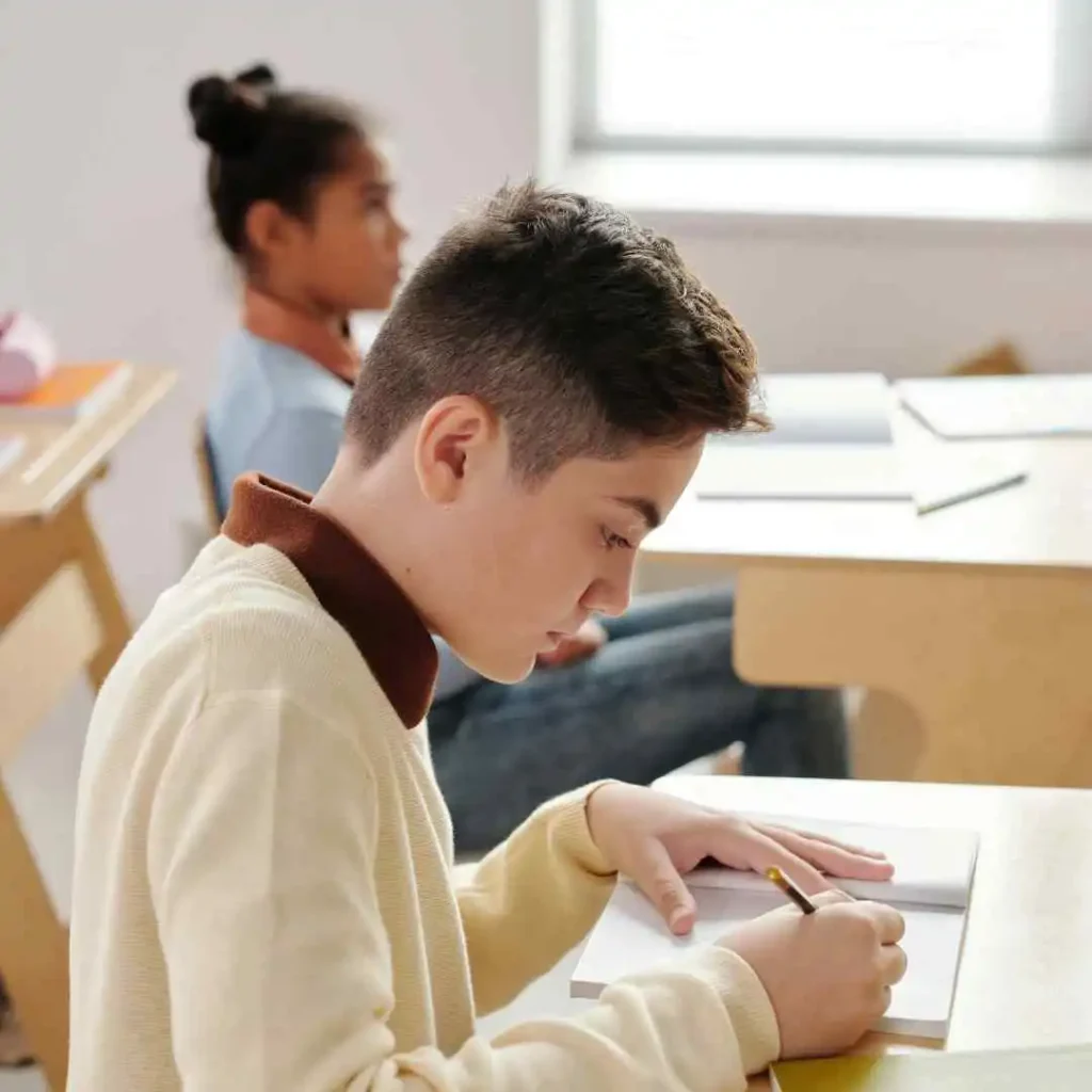 Two students at desks writing, one in a cream sweater, classroom setting.