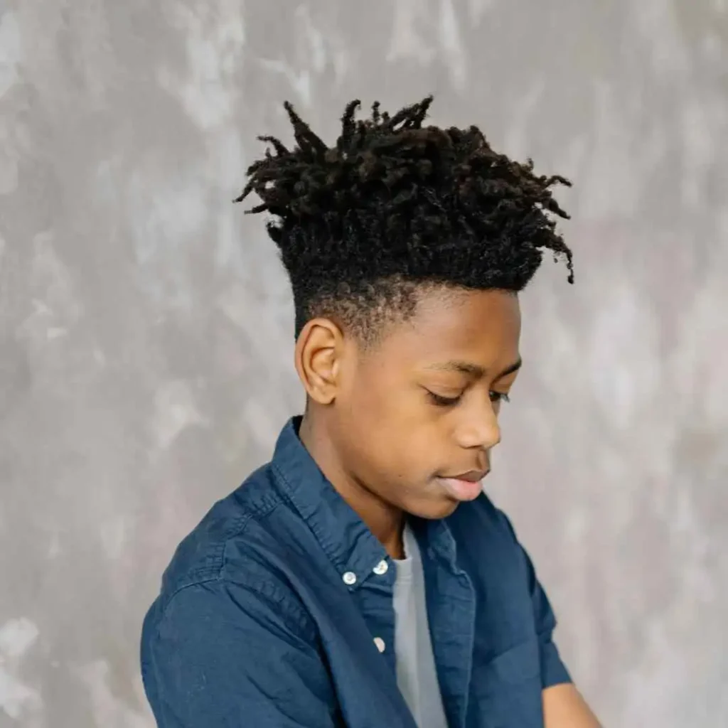 Person with short dreadlocks wearing a denim shirt against a mottled gray background.