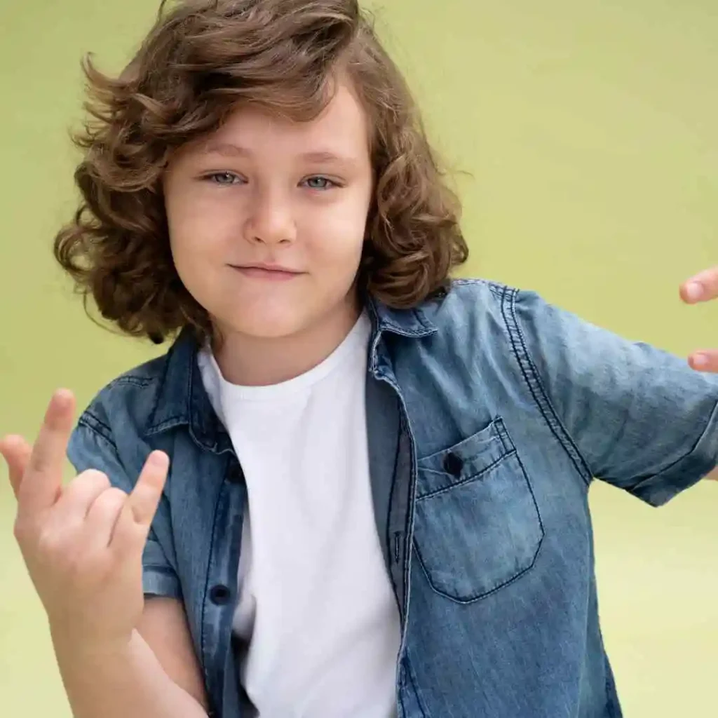 Child in a denim jacket and white shirt with curly hair, gesturing with hands.