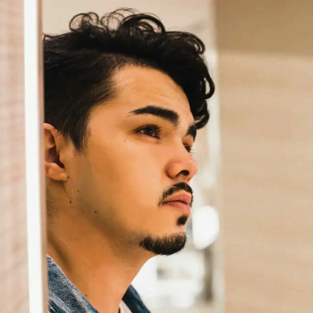 A man with a goatee and a beard looking into a mirror, showcasing haircuts for men with diamond face.