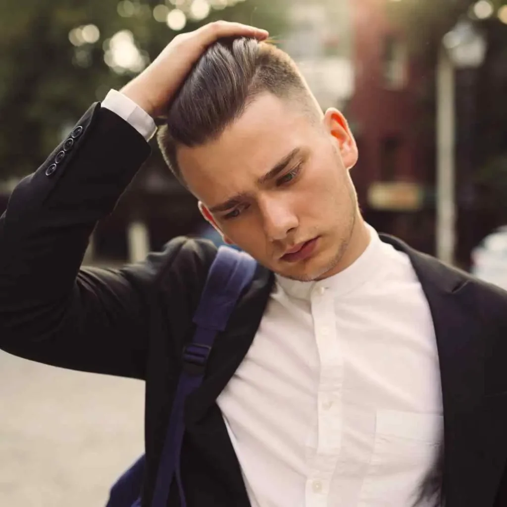 Discover 20 New Pompadour Hairstyles- A Comprehensive Guide 1 A young man dressed in a black suit jacket and white shirt stands outdoors, casually holding his hair styled in pompadour hairstyles while carrying a bag.