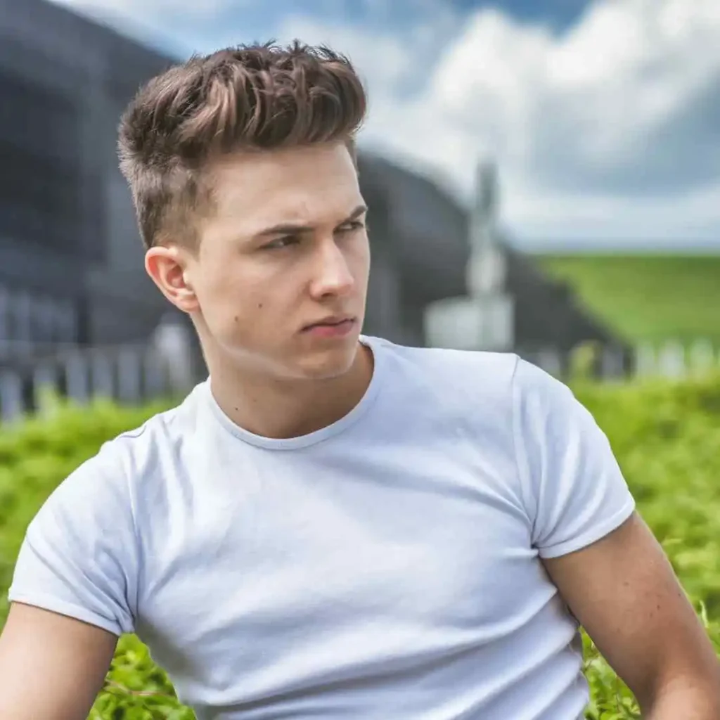 18 Top Men Hairstyles Quiff 1 A young man showcasing MEN HAIRSTYLES QUIFF sits in front of greenery and a modern architectural backdrop, wearing a fitted white t-shirt.