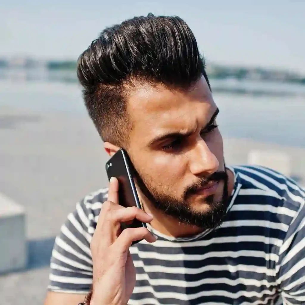A young man showcasing MEN HAIRSTYLES QUIFF, wearing a stylish striped shirt, holding a phone to his ear, standing outdoors by a body of water.