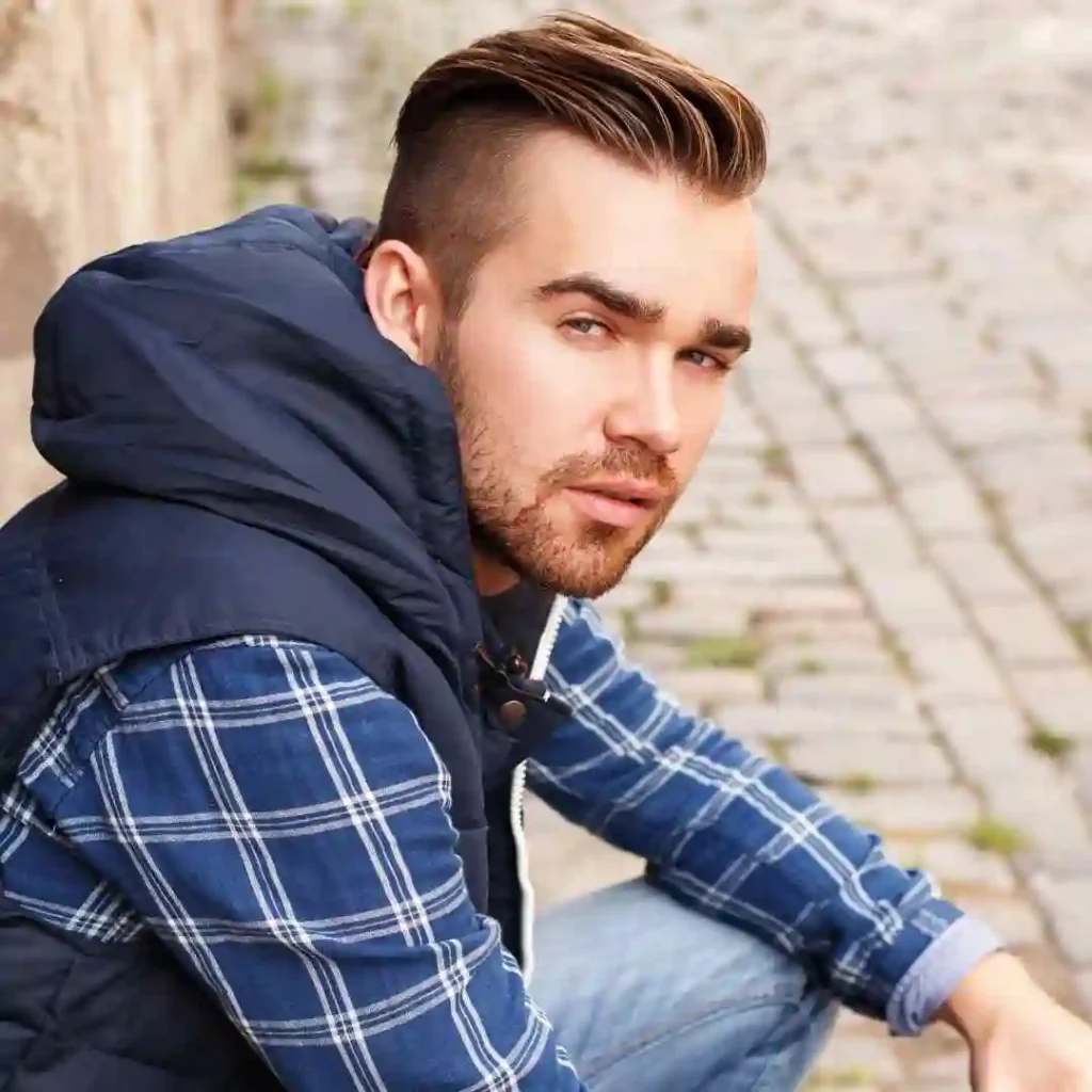A young man sits casually on cobblestone pavement, wearing a plaid shirt under a navy vest, with styled pompadour hairstyles and a relaxed posture.