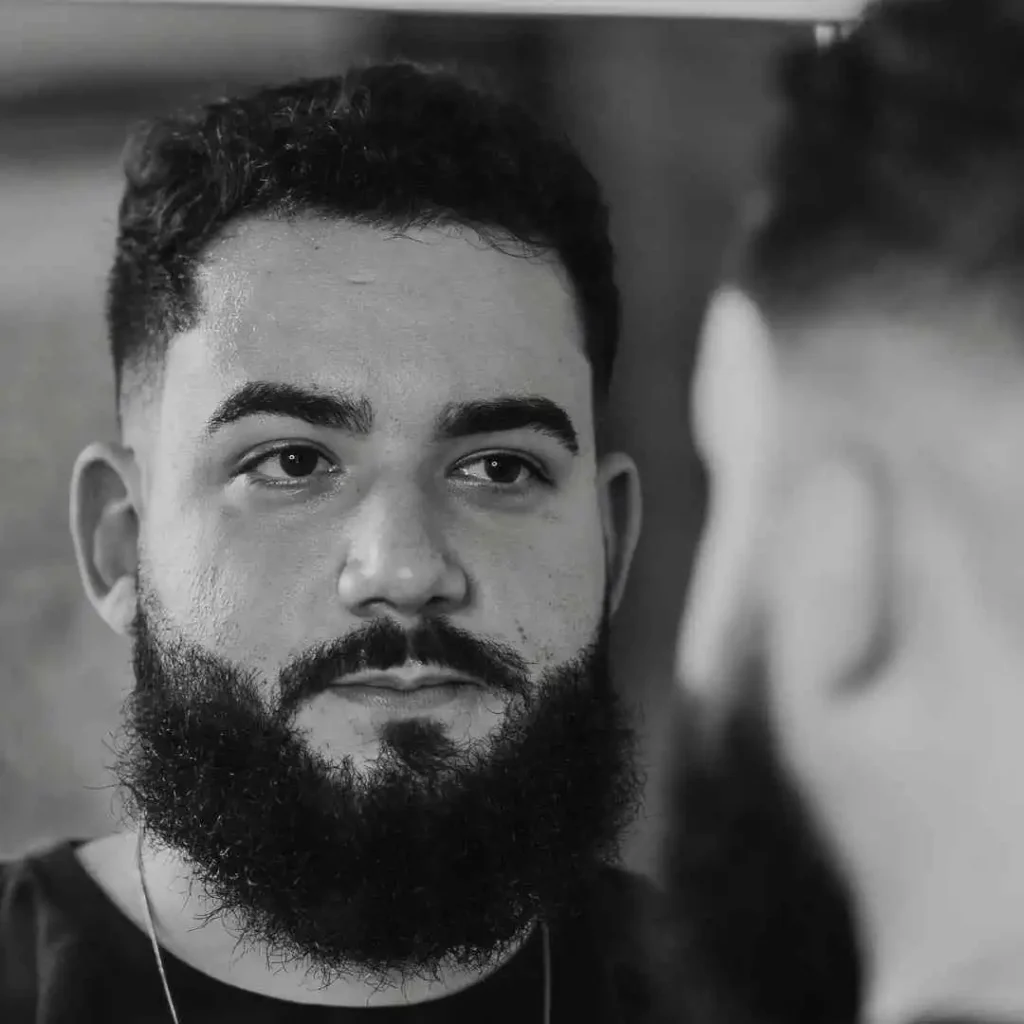 A man with a full beard gazes into a mirror, reflecting a side view of his profile, showcasing one of the best men hairstyles for an oval face. The image is in black and white.