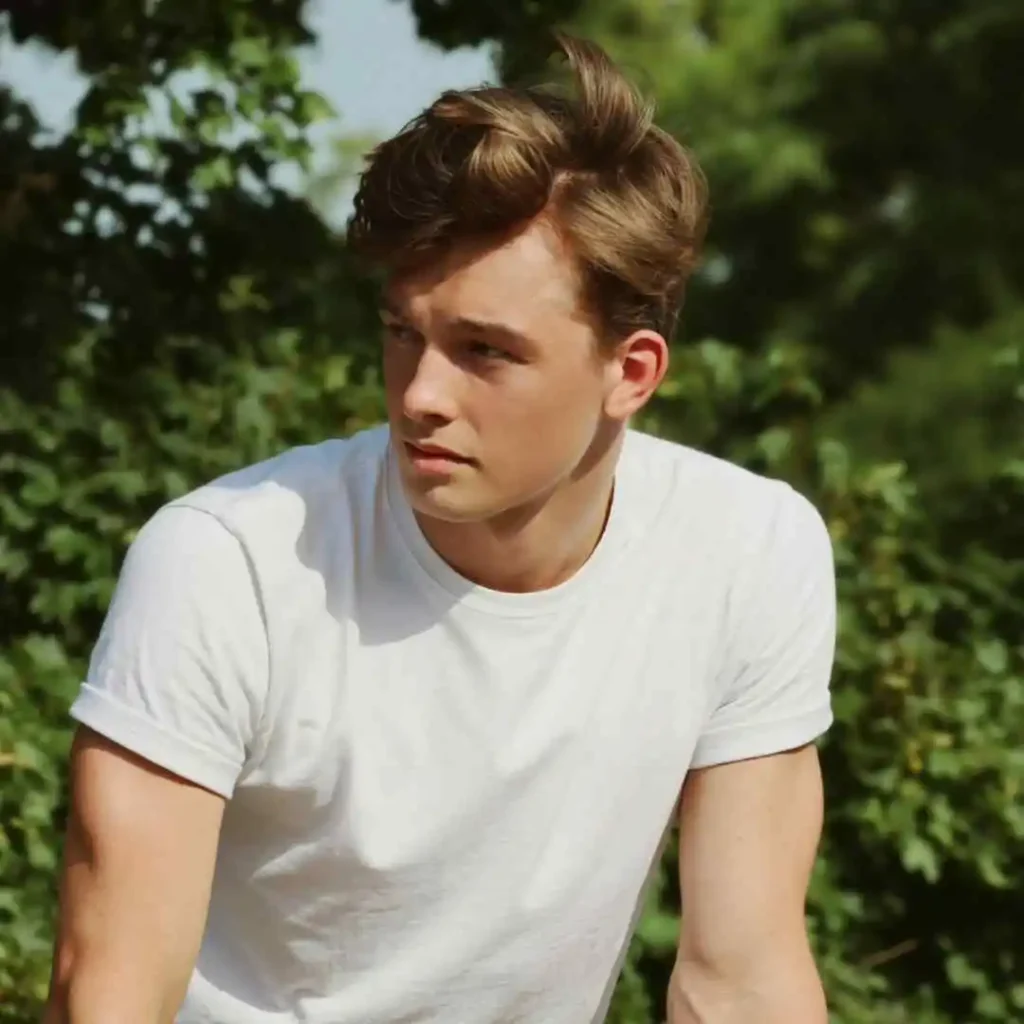 A young man with a stylish MEN HAIRSTYLES QUIFF, wearing a white t-shirt, leans against a picnic table surrounded by lush greenery, enjoying a sunny day outdoors.