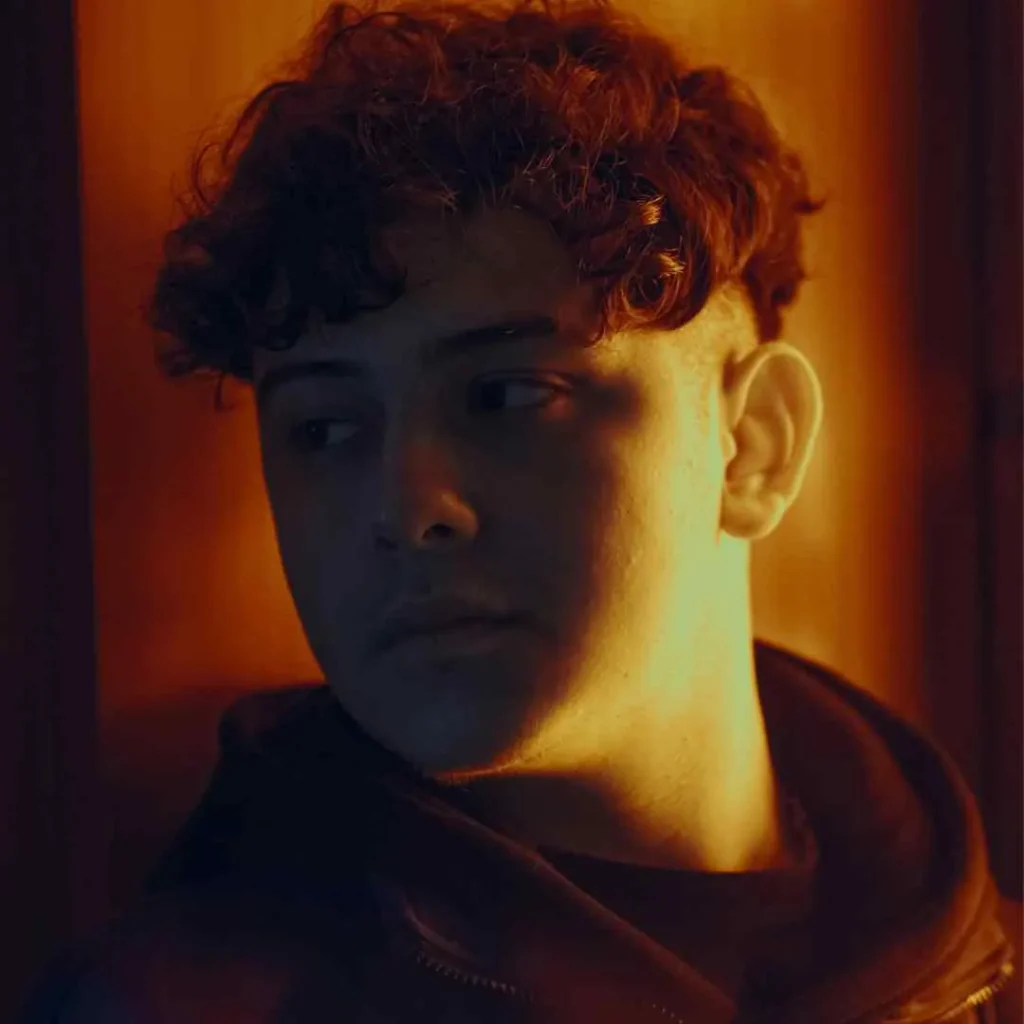 A person with curly hair is seen from the side in a dimly lit room, surrounded by warm orange tones. They wear a black jacket.