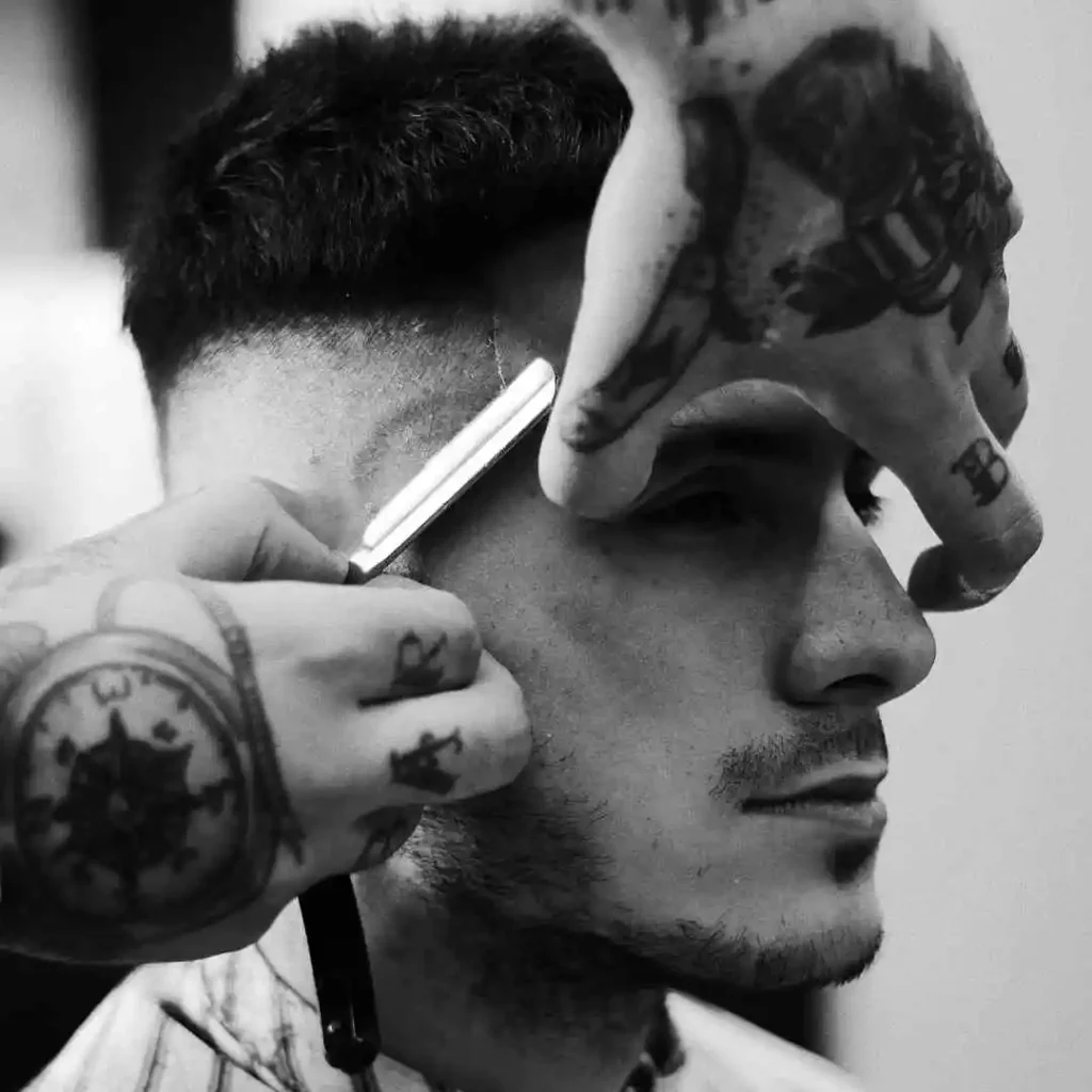 A tattooed barber uses clippers to style the hair of a client in a modern barbershop, showcasing intricate tattoo designs on his arms, while giving a MEN HAIRSTYLES BUZZ CUT.