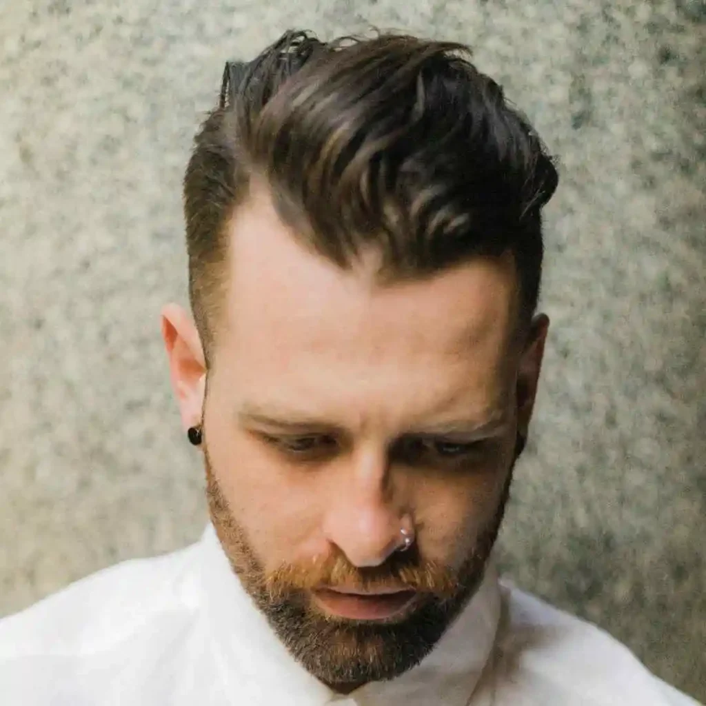 A person with stylish, wavy pompadour hairstyles and earrings, dressed in a white shirt, poses against a textured grey background.