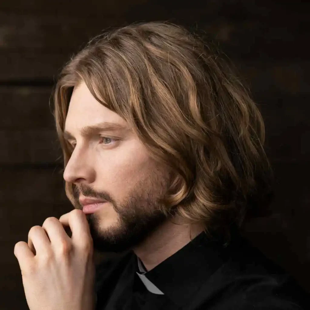 A person with medium-length, wavy blonde hair, showcasing a trendy look for men hairstyles oval face, is dressed in a black collared shirt and posed thoughtfully against a dark background."