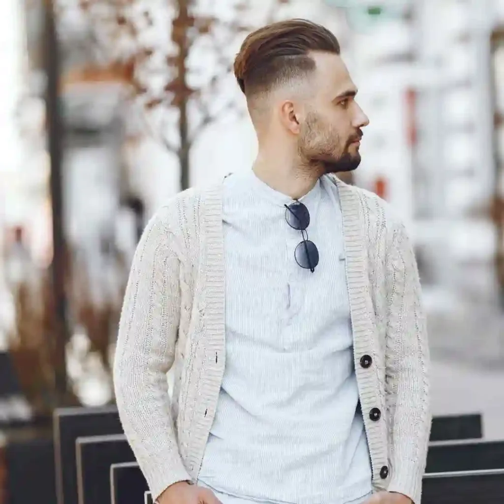 Man in a white shirt and beige cardigan with sunglasses hanging from the neckline.