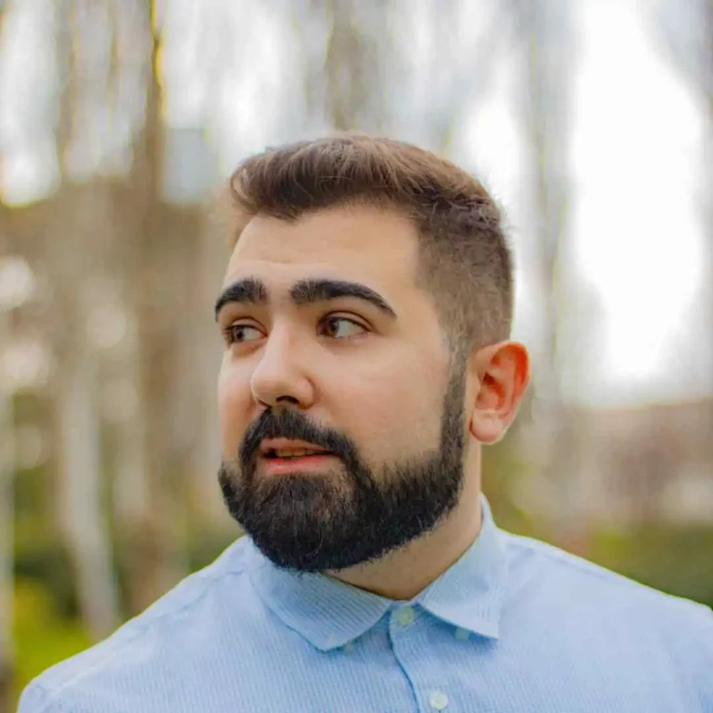 A young man with a buzz cut, representing MEN HAIRSTYLES BUZZ CUT, stands outdoors in a light blue button-up shirt, surrounded by blurred greenery and trees in the background.
