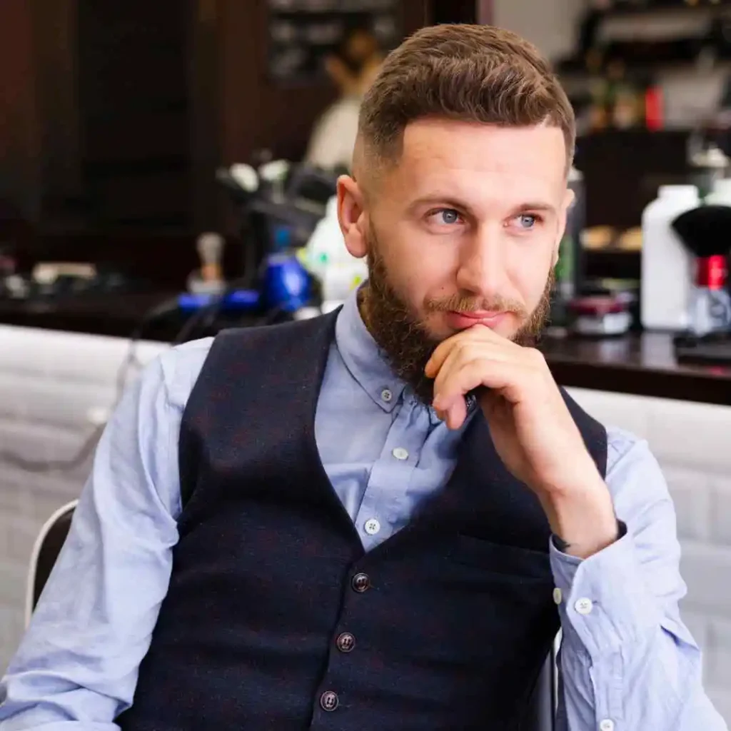 A well-dressed young man in a vest and button-up shirt sits thoughtfully in a barbershop, surrounded by grooming tools, showcasing modern men short hairstyles.