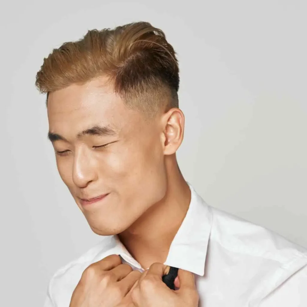 A young person with stylish, blonde MEN HAIRSTYLES QUIFF adjusts their white shirt collar against a simple gray background.