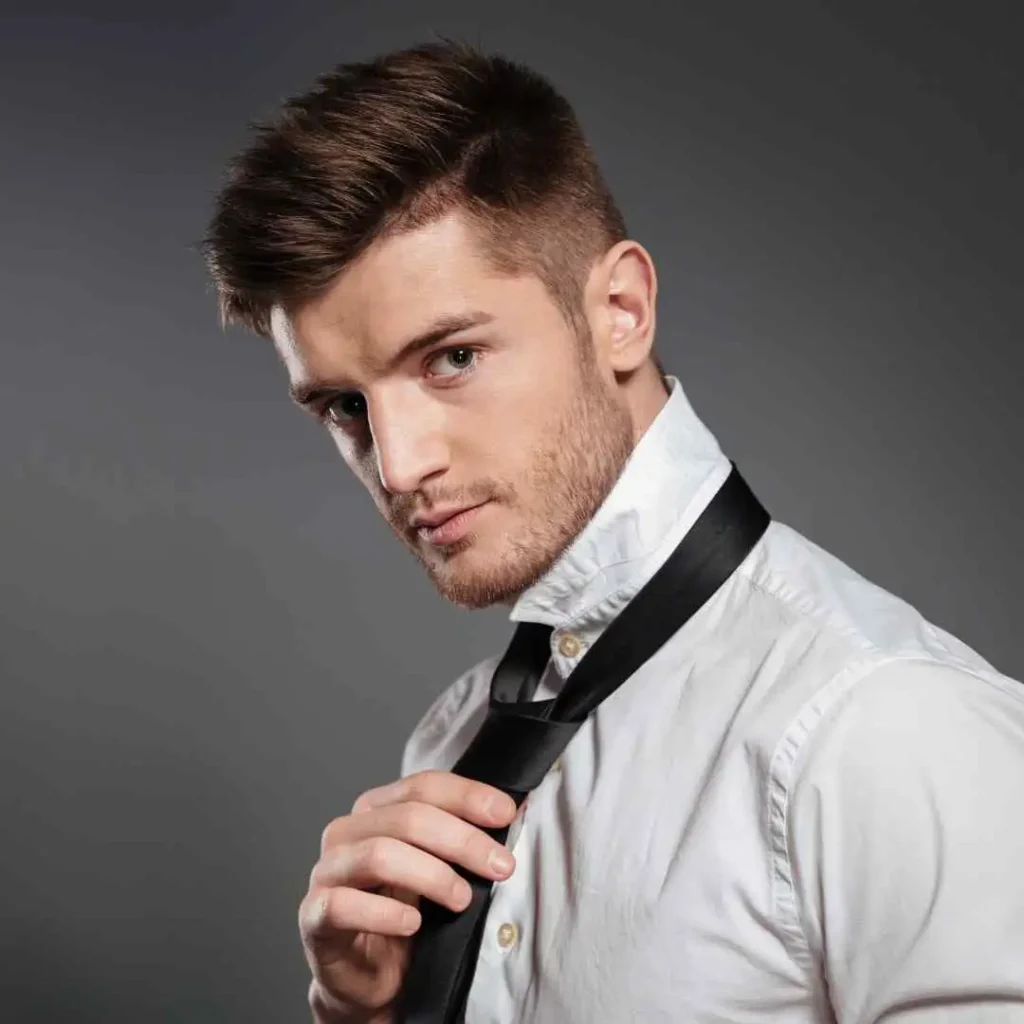 A man with classic POMPADOUR HAIRSTYLES in a white shirt meticulously adjusts a black tie against a grey background, exuding a formal and polished vibe.