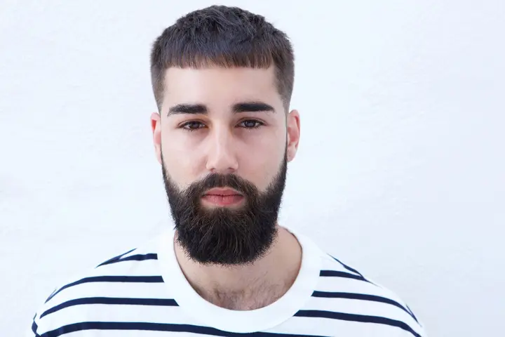 A man with a short beard and dark hair wears a white and navy striped shirt, standing against a plain white background, showcasing a men’s hairstyles buzz cut.