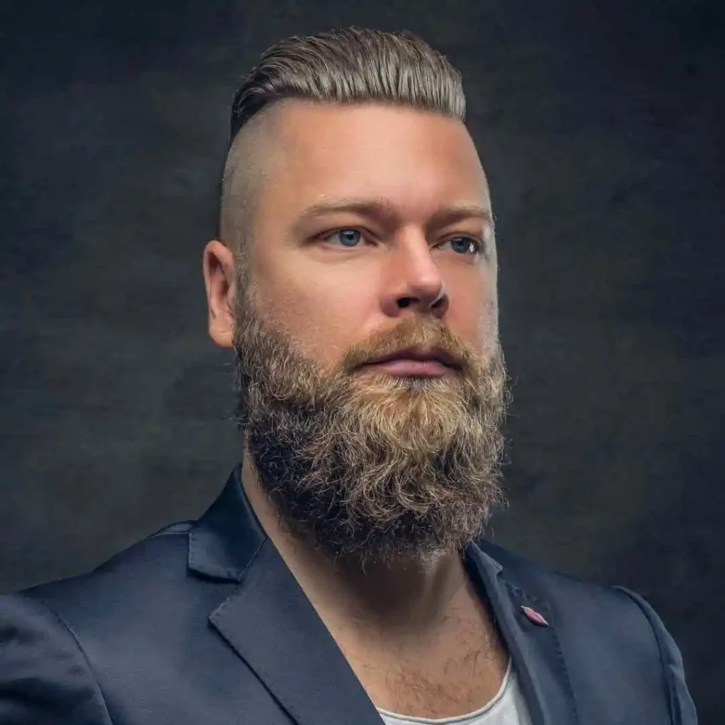 A man with a thick beard and styled pompadour hairstyles wears a dark blazer over a light shirt, set against a dark background.