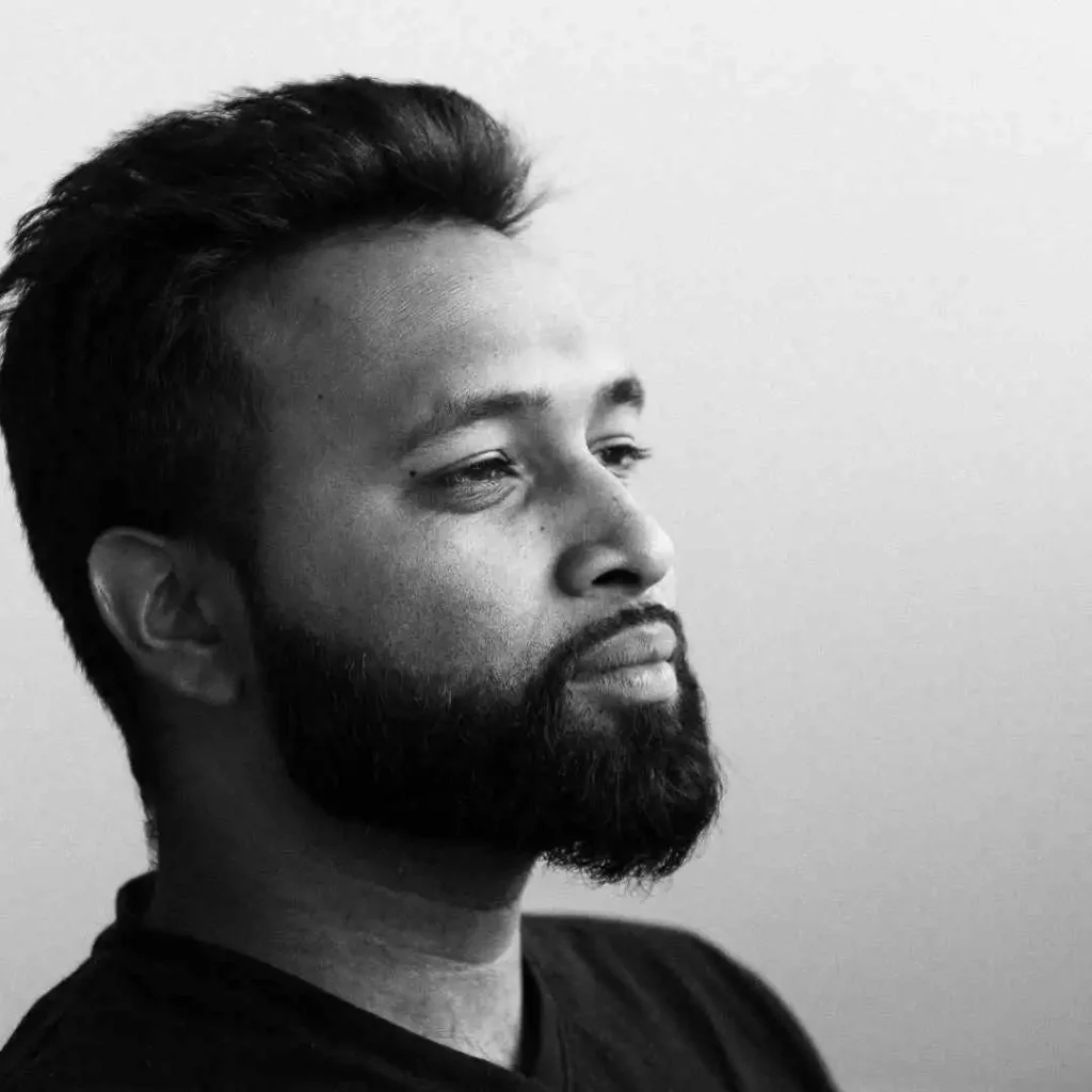 A man with dark hair and a beard is seen in profile, wearing a black t-shirt, showcasing a stylish MEN HAIRSTYLES QUIFF against a soft gray background.