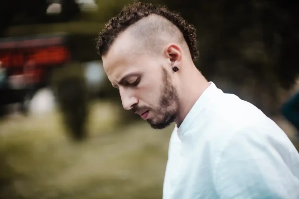 A person with a MEN HAIRSTYLES BUZZ CUT, curly hair on top, and an earring wears a white shirt, looking down in a natural outdoor setting.