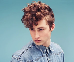 A person with men's short curly haircuts, wearing a denim shirt, poses against a light blue background.