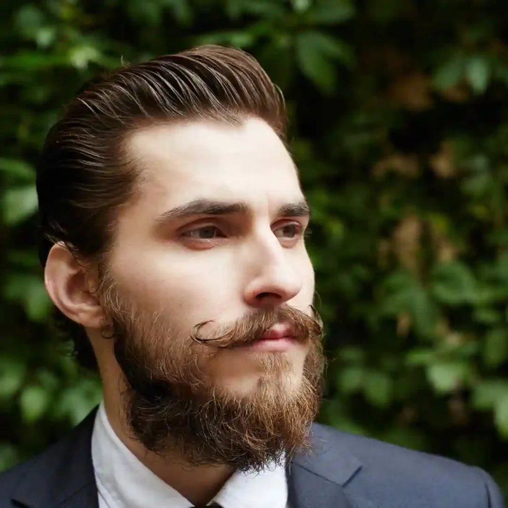 A man with styled hair and a beard stands against a backdrop of lush green foliage, dressed in a formal suit and tie, showcasing modern men short hairstyles.