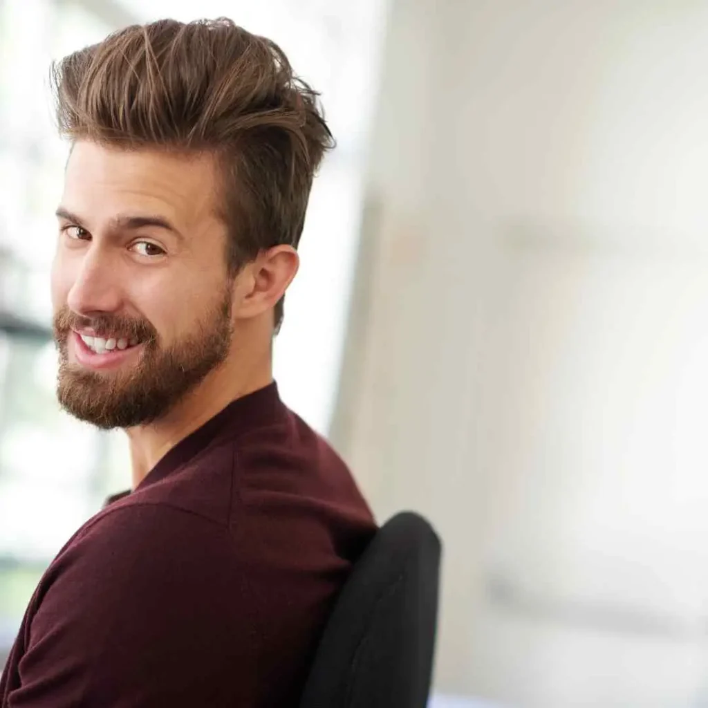 A young man showcasing MEN HAIRSTYLES QUIFF with stylish, tousled hair sits sideways in a modern office environment, wearing a maroon shirt.