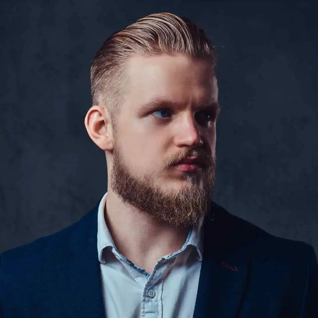 A well-groomed man with a beard and styled pompadour hairstyle, wearing a dark suit jacket over a light blue shirt, set against a dark background.