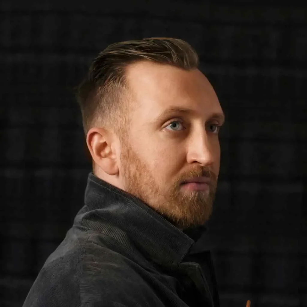 A man with short brown hair wearing a black coat, side-profile against a dark background, holds an object in his right hand, showcasing a stylish men’s hairstyles buzz cut.