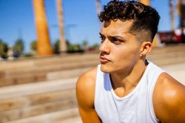 A young man with MEN'S SHORT CURLY HAIRCUTS, wearing a sleeveless white shirt, sitting on wooden steps and enjoying a sunny day in an outdoor setting.