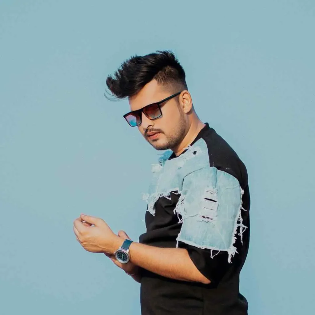 A person with pompadour hairstyles, stylish hair, and glasses wears a torn denim shirt against a clear blue backdrop, showcasing a casual pose.