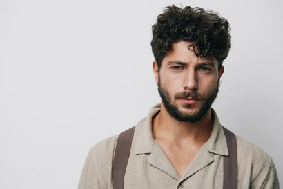 A young man with men's short curly haircuts, wearing a beige button-up shirt and suspenders, posing against a plain white background.