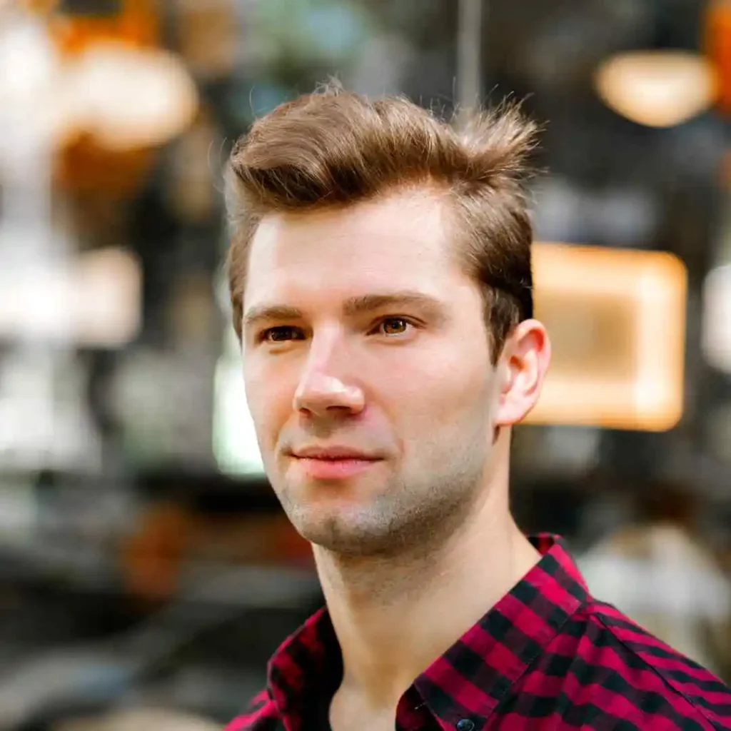 A young man with styled brown hair, showcasing a pompadour hairstyle, wearing a black and red checked shirt stands against a blurred urban background.
