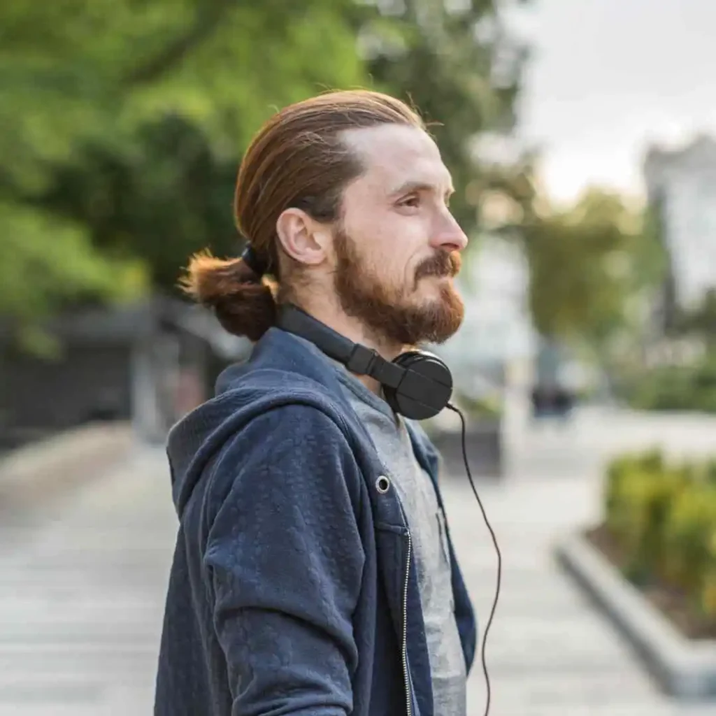 A person showcasing a trendy men hairstyle for an oval face, with long hair tied back, wearing headphones, standing outdoors in a park setting. Trees and pathways are visible in the background.