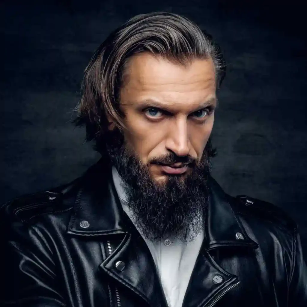 A man with long, wavy hair styled in a pompadour hairstyle and a thick beard wears a black leather jacket over a white shirt, posed against a dark backdrop.