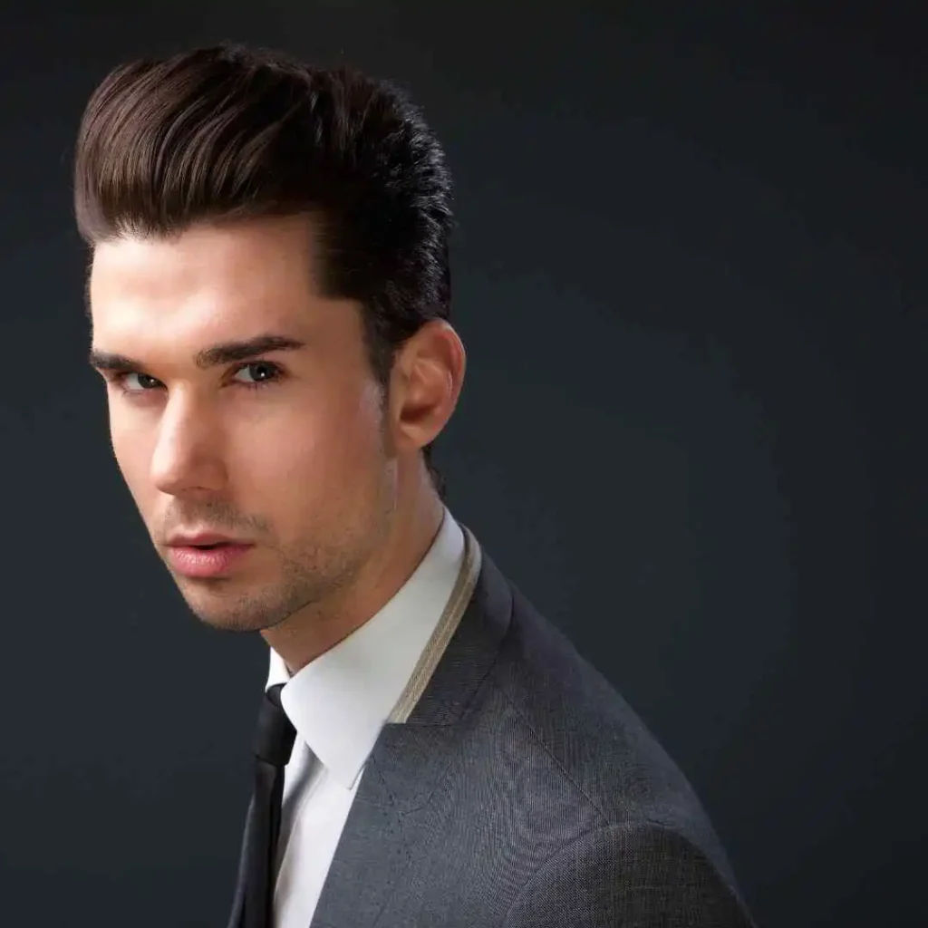 A young man showcasing MEN HAIRSTYLES QUIFF with stylish, slicked-back hair, wearing a tailored suit and tie, set against a dark, neutral background.