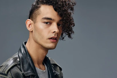 A person showcasing MEN'S SHORT CURLY HAIRCUTS, wearing a black leather jacket and a gray t-shirt against a gray background.