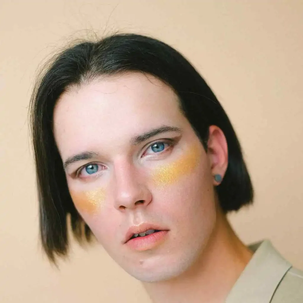 A person with sleek black hair, wearing a beige collared shirt, stands against a soft peach background, exuding a calm vibe, showcasing one of the best men hairstyles for an oval face.