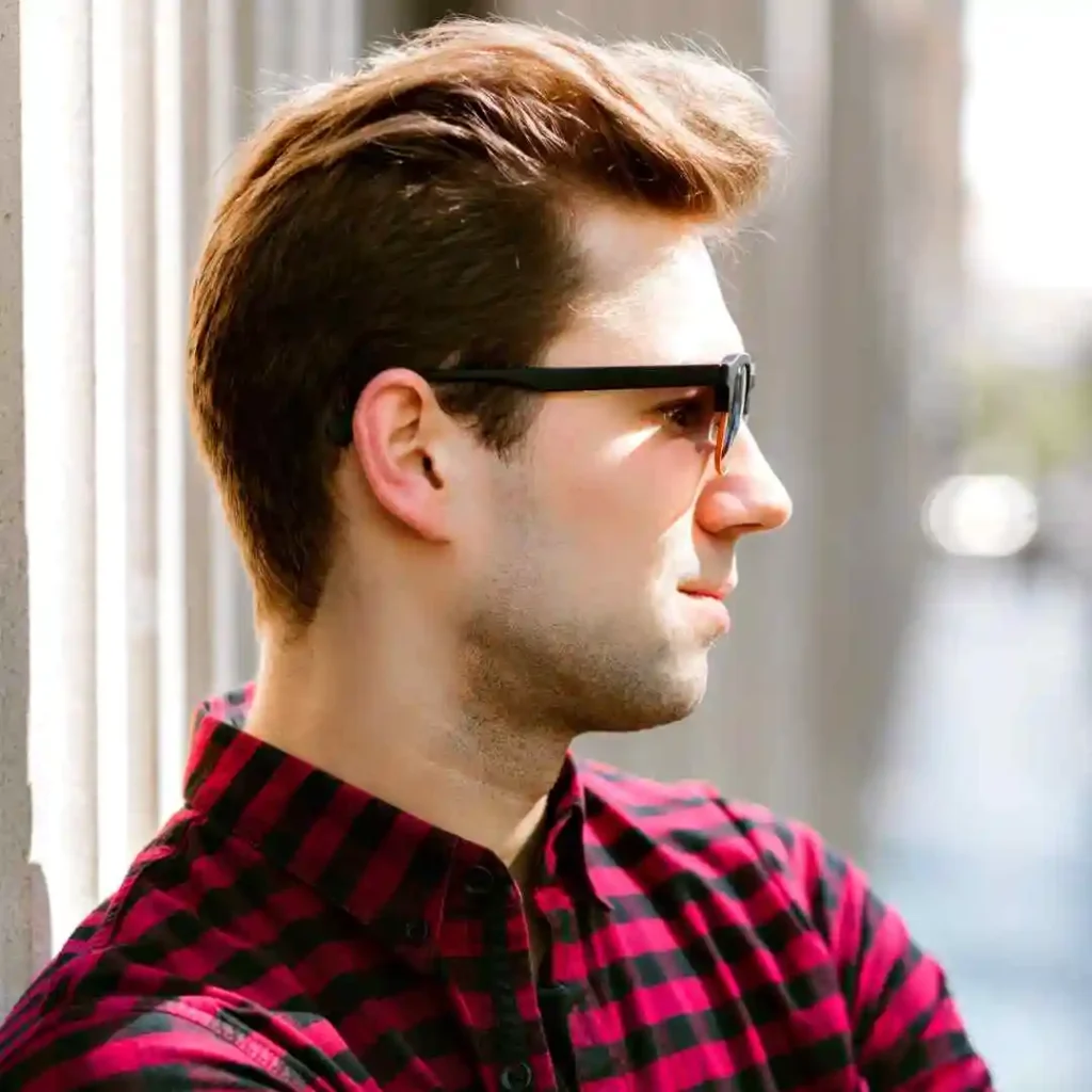 A young man with stylishly tousled hair, sporting a Pompadour hairstyle, leans against a column, wearing a red and black plaid shirt, in an urban setting