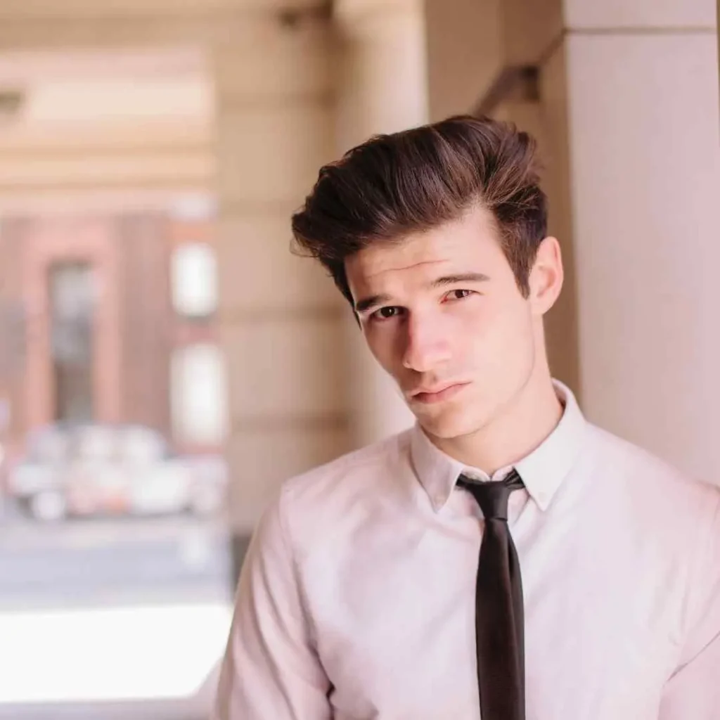 "A young man stands in a softly lit corridor, wearing a light shirt and a dark tie, showcasing MEN HAIRSTYLES QUIFF with a stylish, modern look.