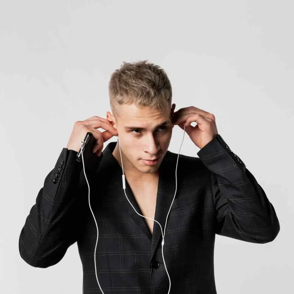 A person in a stylish black suit adjusts earbuds while holding a phone, set against a plain gray background, showcasing modern men short hairstyles.