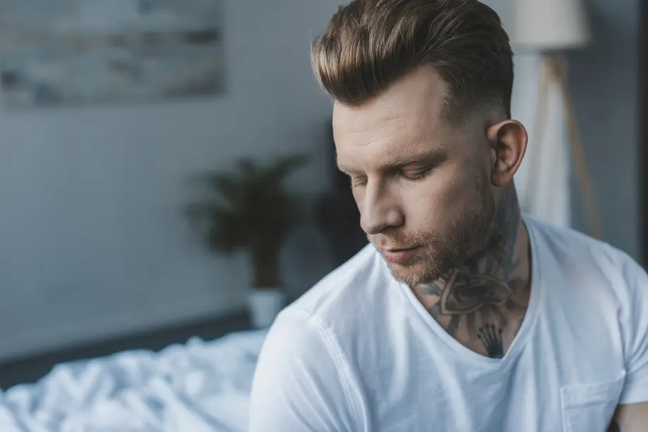 A young man with styled hair in a pompadour hairstyle and visible tattoos wears a white t-shirt, sitting on a bed in a softly lit, modern room.