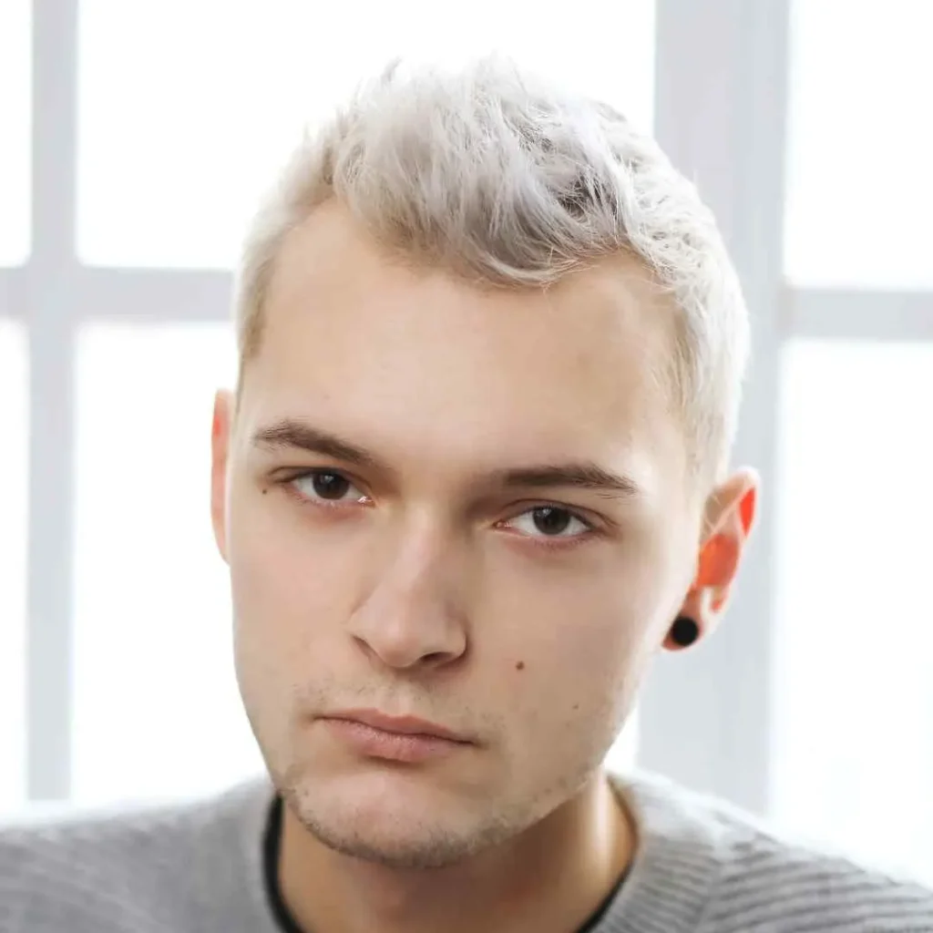 Discover 20 New Pompadour Hairstyles- A Comprehensive Guide 2 A person with short, light blonde hair styled in pompadour hairstyles is sitting indoors, framed by soft natural light coming from large windows.