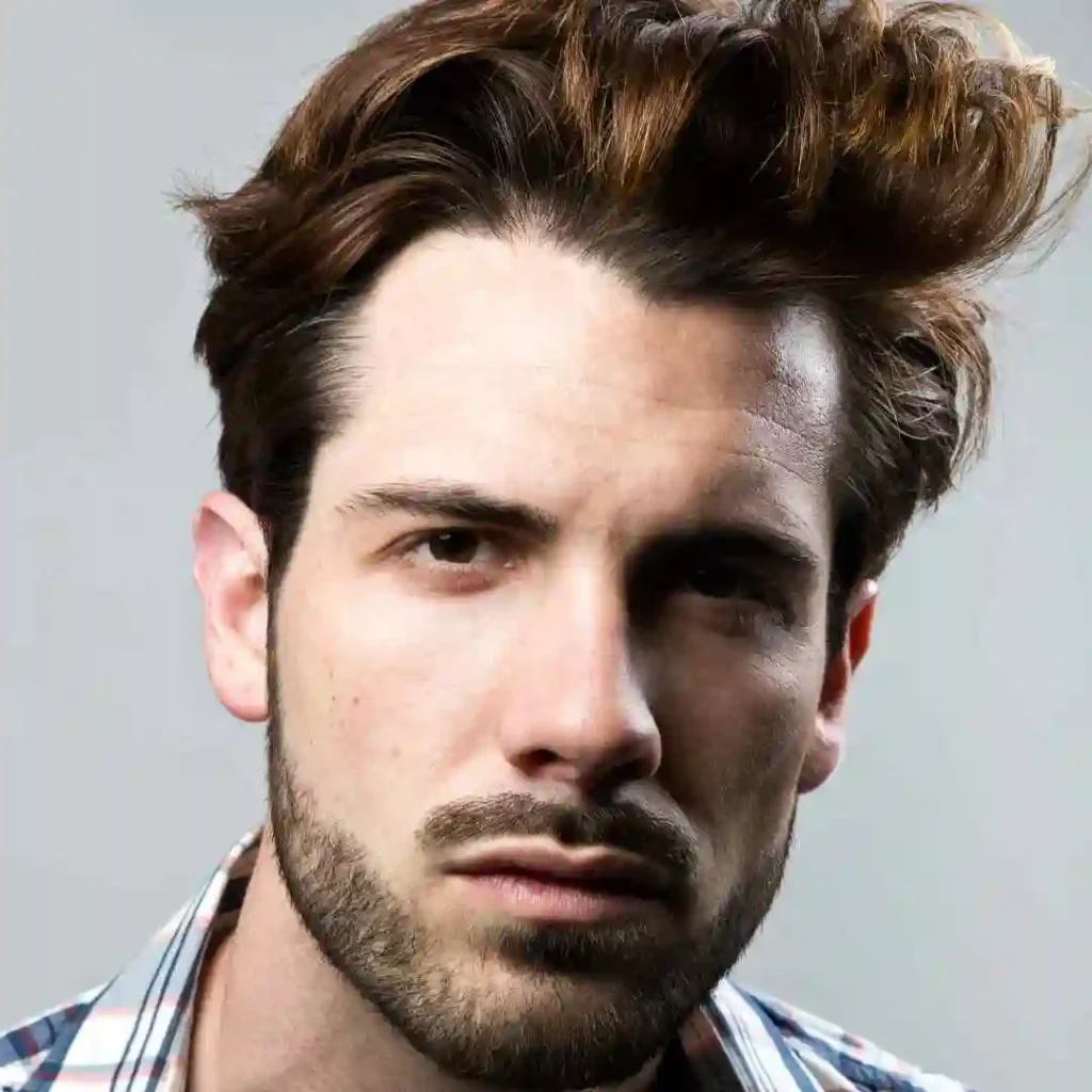 18 Top Men Hairstyles Quiff 2 A person with tousled, wavy hair styled in a MEN HAIRSTYLES QUIFF, wearing a plaid shirt, standing against a neutral gray background.