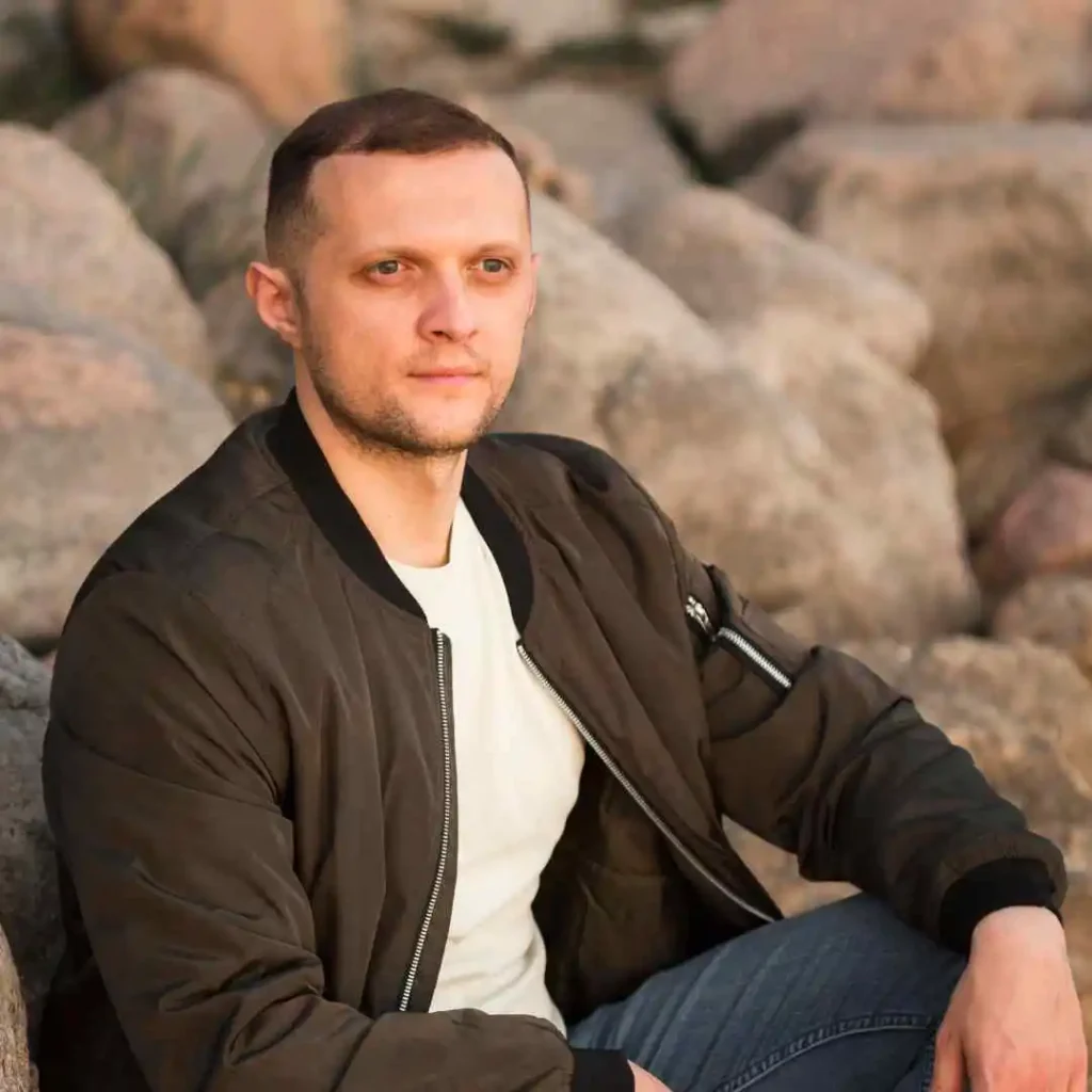 Modern Men Short Hairstyles- A Comprehensive Guide 2 A person with MODERN MEN SHORT HAIRSTYLES sitting on large rocks, wearing a black bomber jacket and white shirt, with legs crossed, in a natural outdoor setting.