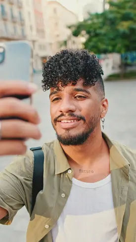 A person with men's short curly haircuts takes a selfie outdoors, wearing a light jacket and showcasing a tattoo on their neck.