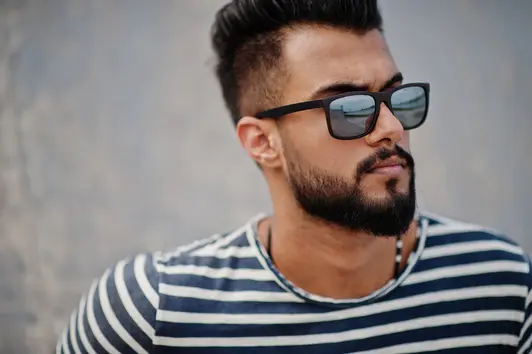 "A man with sunglasses and a stylish beard showcases POMPADOUR HAIRSTYLES against a textured gray wall, wearing a striped shirt.