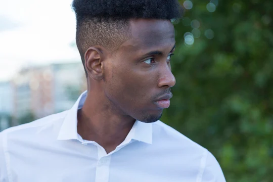 A young man in a white shirt stands outdoors, with greenery and a blurred urban background. His hairstyle features MEN'S SHORT CURLY HAIRCUTS, looking both modern and stylish.