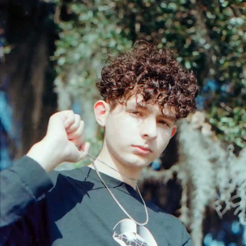 A person with a men's short curly haircut wearing a black sweatshirt, holding a chain with a pendant, standing outdoors with lush greenery in the background.