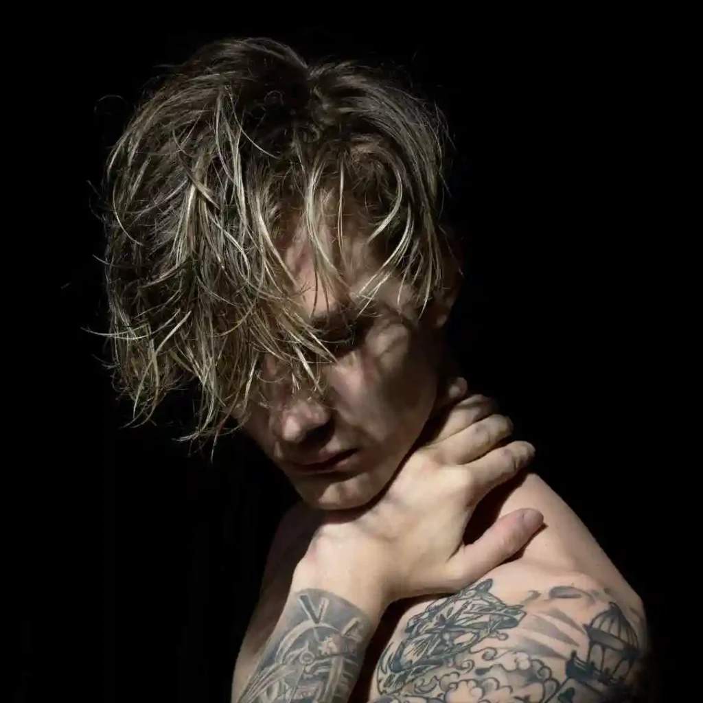 A person with tousled hair and intricate tattoos on their arms poses thoughtfully against a dark background.